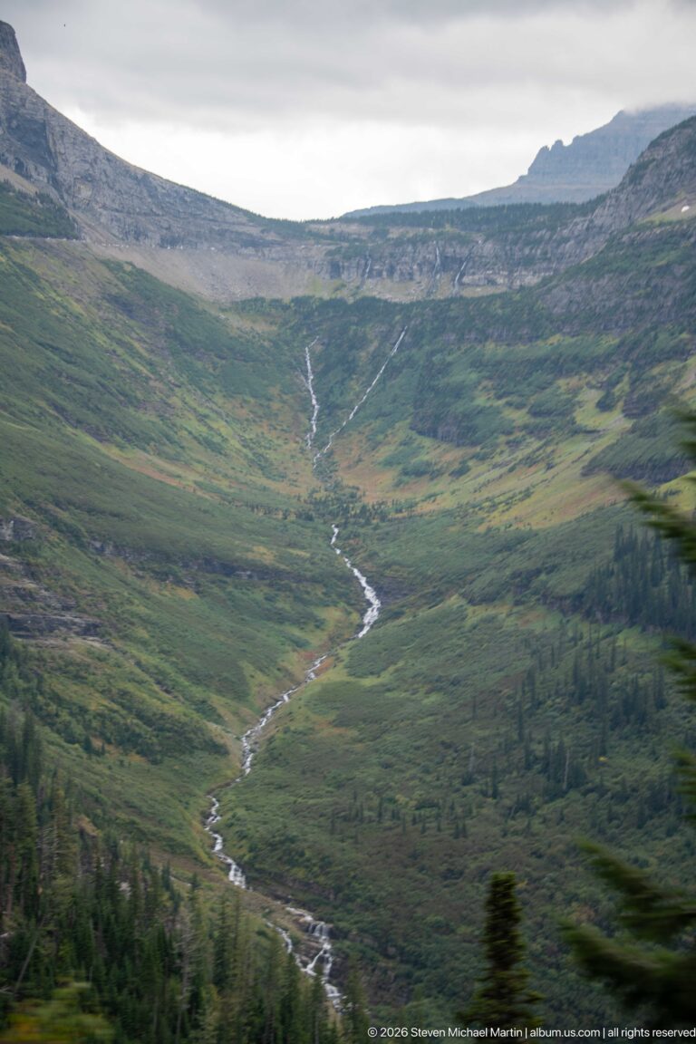 Narrow winding stream descending through a broad green alpine basin below layered rocky slopes.