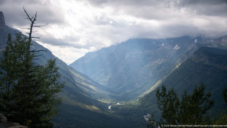 Winding bright stream on the floor of a deep forested valley, framed by trees beneath low storm clouds.