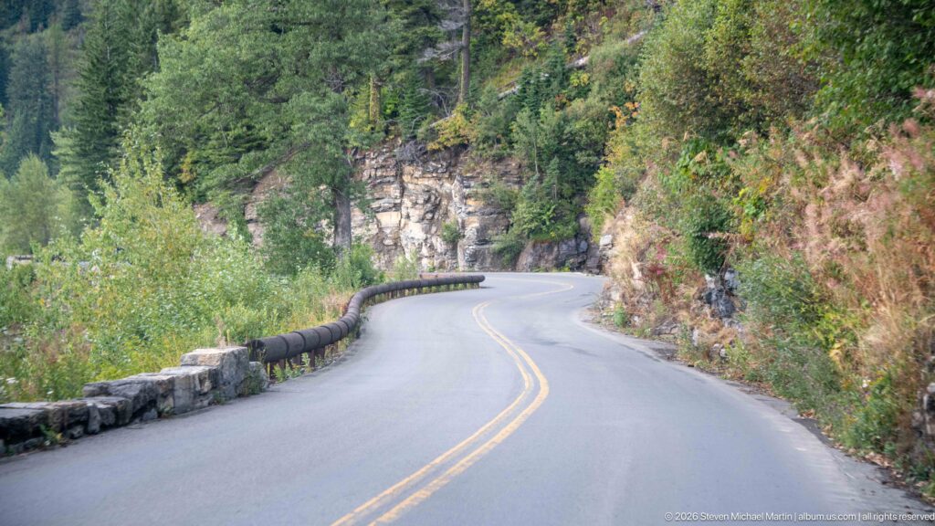 Two-lane Going-to-the-Sun Road curving beside a stone guardwall, dense vegetation, and a rocky mountainside.