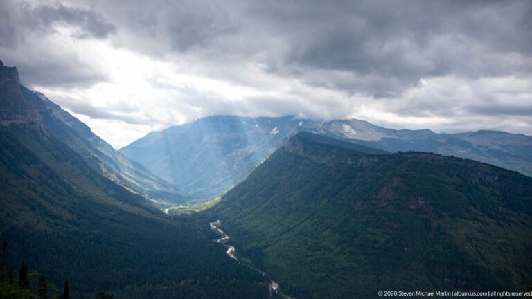Elevated view of a winding stream in a forested mountain valley beneath dark clouds and a broad shaft of sunlight.