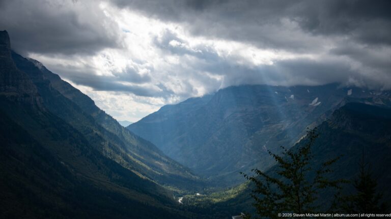 Sunbeams pierce dark clouds above a steep forested mountain valley, illuminating patches of the valley floor.