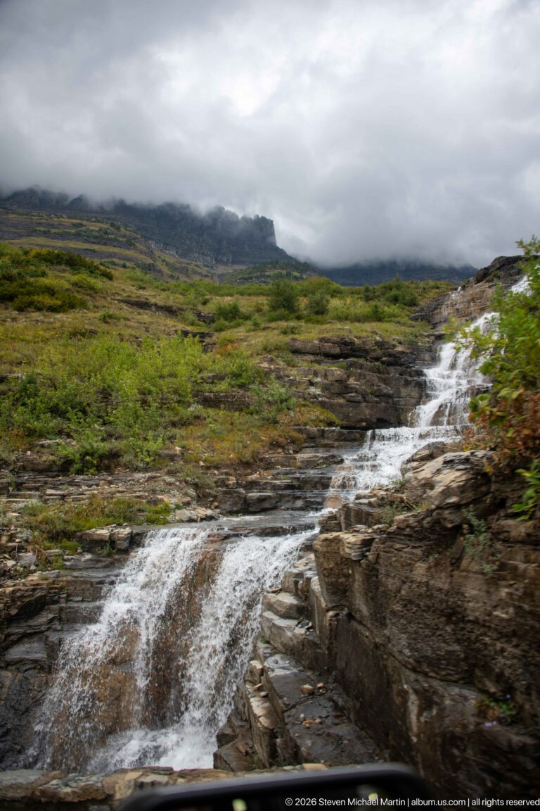 Small cascade spilling over dark rock ledges beside low alpine vegetation, with a cloud-covered slope beyond.