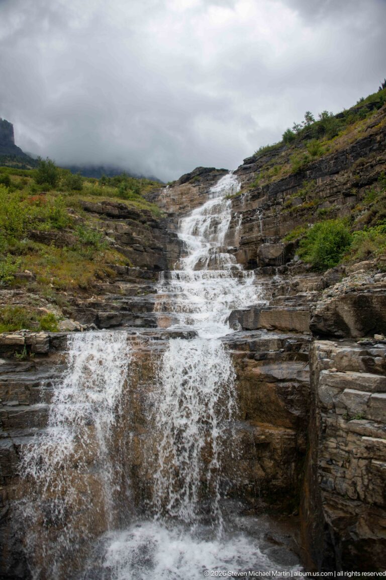 Stepped mountain cascade flowing across broad rock shelves surrounded by low green vegetation under gray clouds.