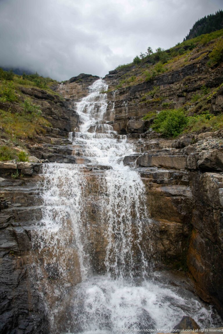 Broad white cascade flowing over layered brown bedrock amid low alpine vegetation beneath an overcast sky.