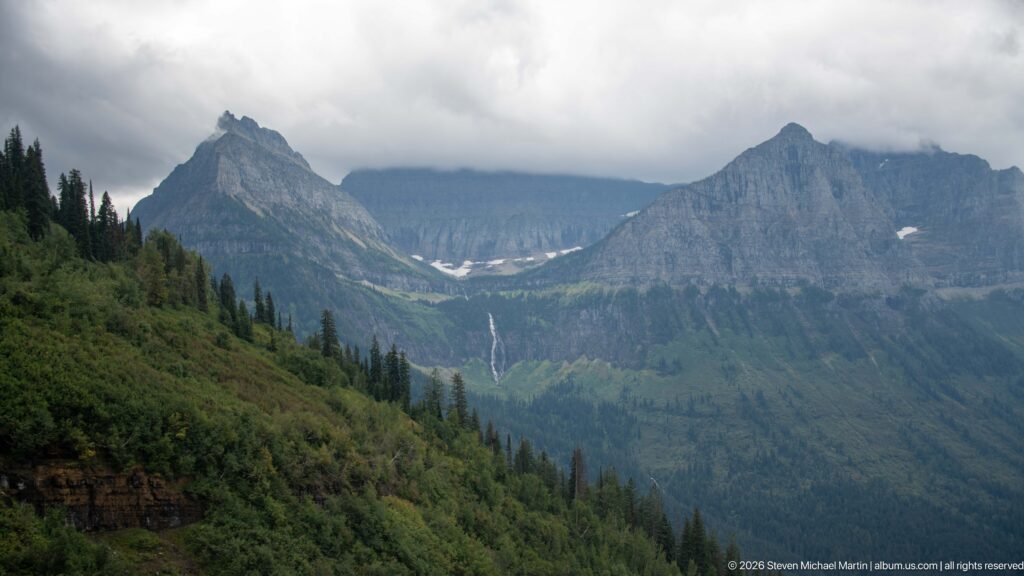 Jagged mountain peaks and a forested glacial valley beneath low clouds, with small snowfields and a distant waterfall.