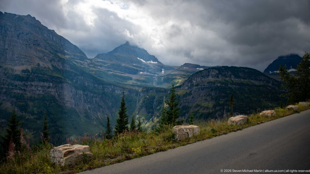 Storm clouds hang over rugged layered peaks with conifers, roadside stone blocks, and small snow patches.