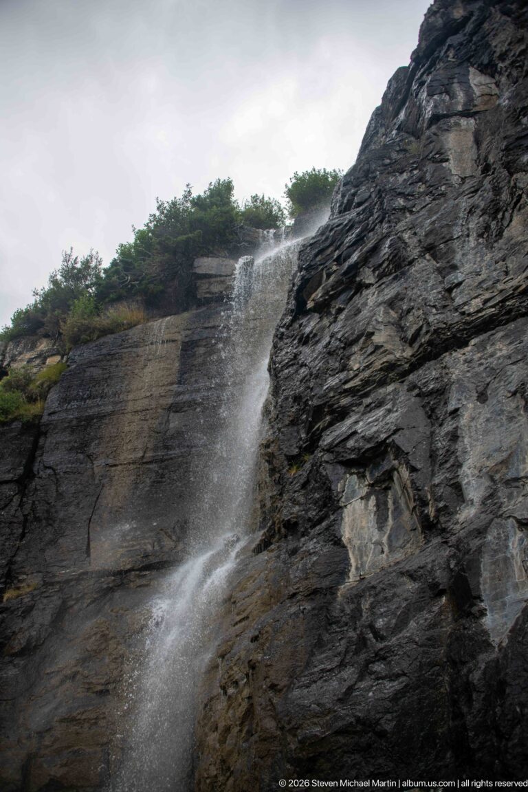 Waterfall spills from a vegetated ledge over dark fractured rock and disperses into mist.