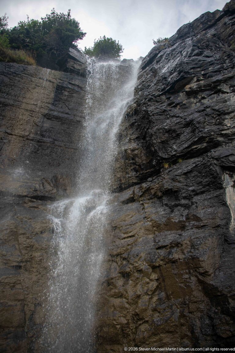 Thin waterfall blows into spray as it falls over a dark layered rock cliff.