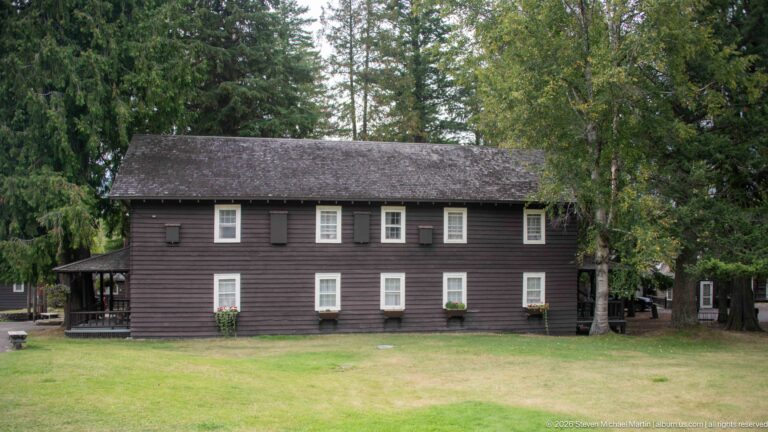 Long two-story dark wooden building with evenly spaced white-trimmed windows, shingled roof, porch, and lawn.