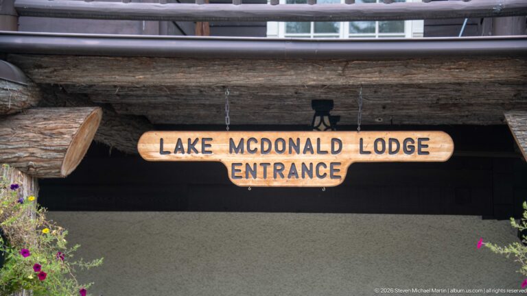 Wooden Lake McDonald Lodge Entrance sign hanging beneath a large log beam, with dark siding and flowers nearby.