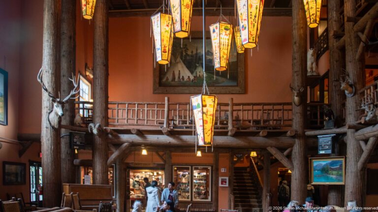 Tall lodge interior with painted lanterns, log columns, upper gallery, mounted wildlife, staircase, and visitors below.