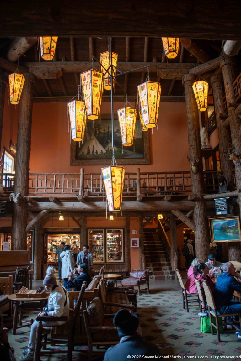 Visitors seated in the spacious lodge lobby beneath painted lanterns, with log gallery, staircase, display cases, and patterned carpet.