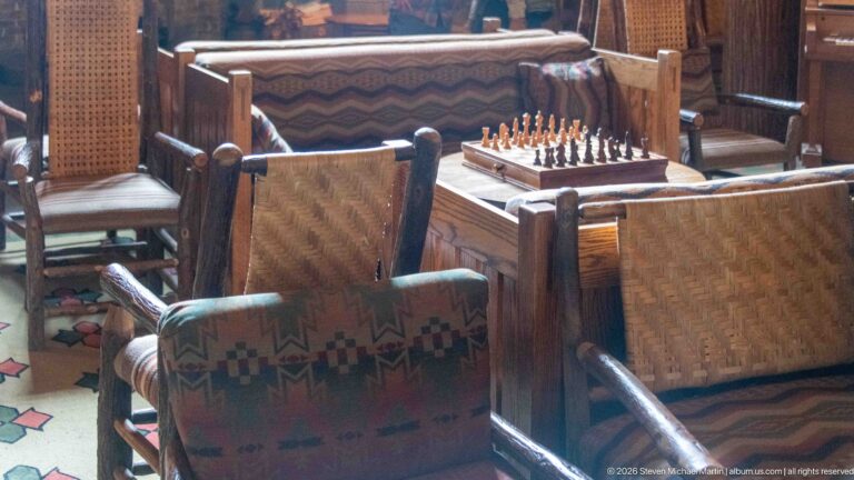 Chess set on a small table surrounded by rustic wooden chairs with woven backs and patterned upholstery.