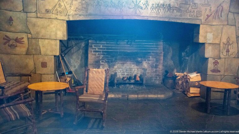 Broad fireplace with pictographic decoration on its masonry surround, flanked by rustic chairs and small wooden tables.