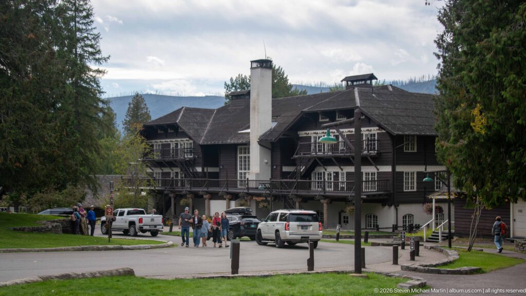 Lake McDonald Lodge beyond a drive with visitors and vehicles, showing balconies, exterior stairs, chimney, and forested hills behind.