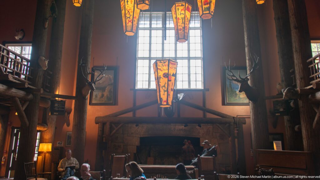 Tall lodge lobby with stone fireplace, painted hanging lantern, log columns, mounted wildlife, upper gallery, and visitors.