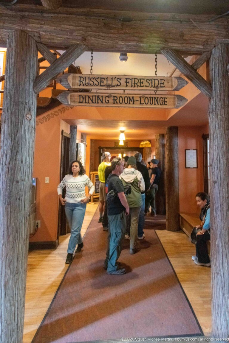 Log-framed corridor inside Lake McDonald Lodge with hanging signs for Russell’s Fireside and the dining room and lounge, with visitors beyond.