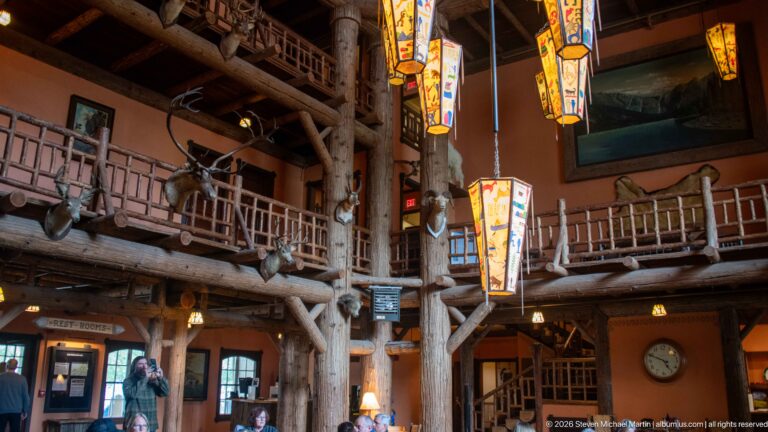 Two-story Lake McDonald Lodge lobby with massive log columns, rustic balcony railings, colorful hanging lanterns, and visitors on the galleries.