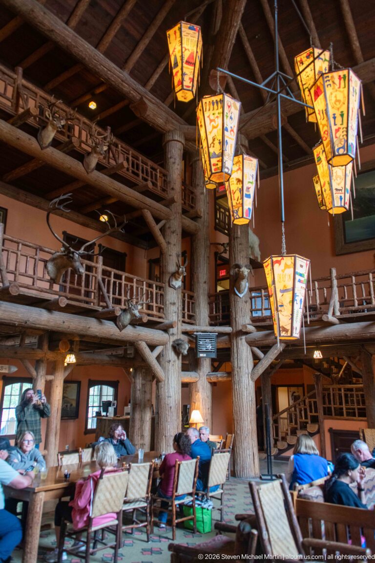 Lake McDonald Lodge lobby with seated visitors beneath colorful hanging lanterns, massive log columns, and rustic upper galleries.