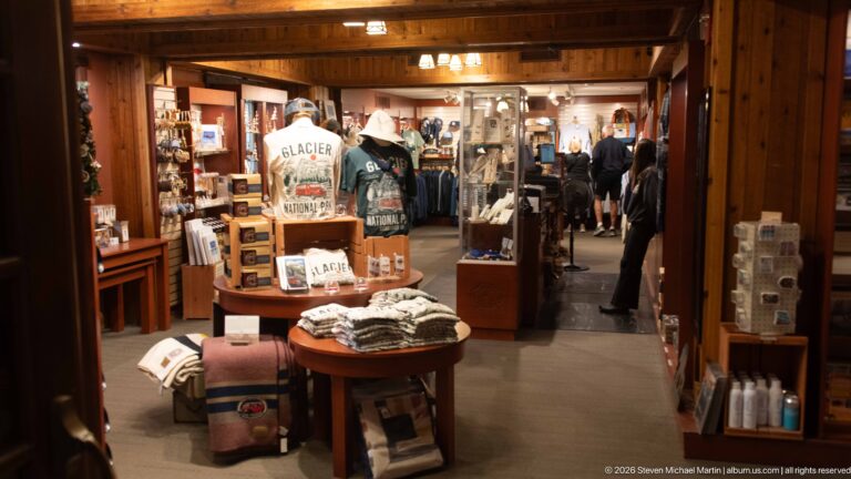 Wood-paneled gift shop inside Lake McDonald Lodge with tables and displays of Glacier apparel, books, souvenirs, and visitor merchandise.