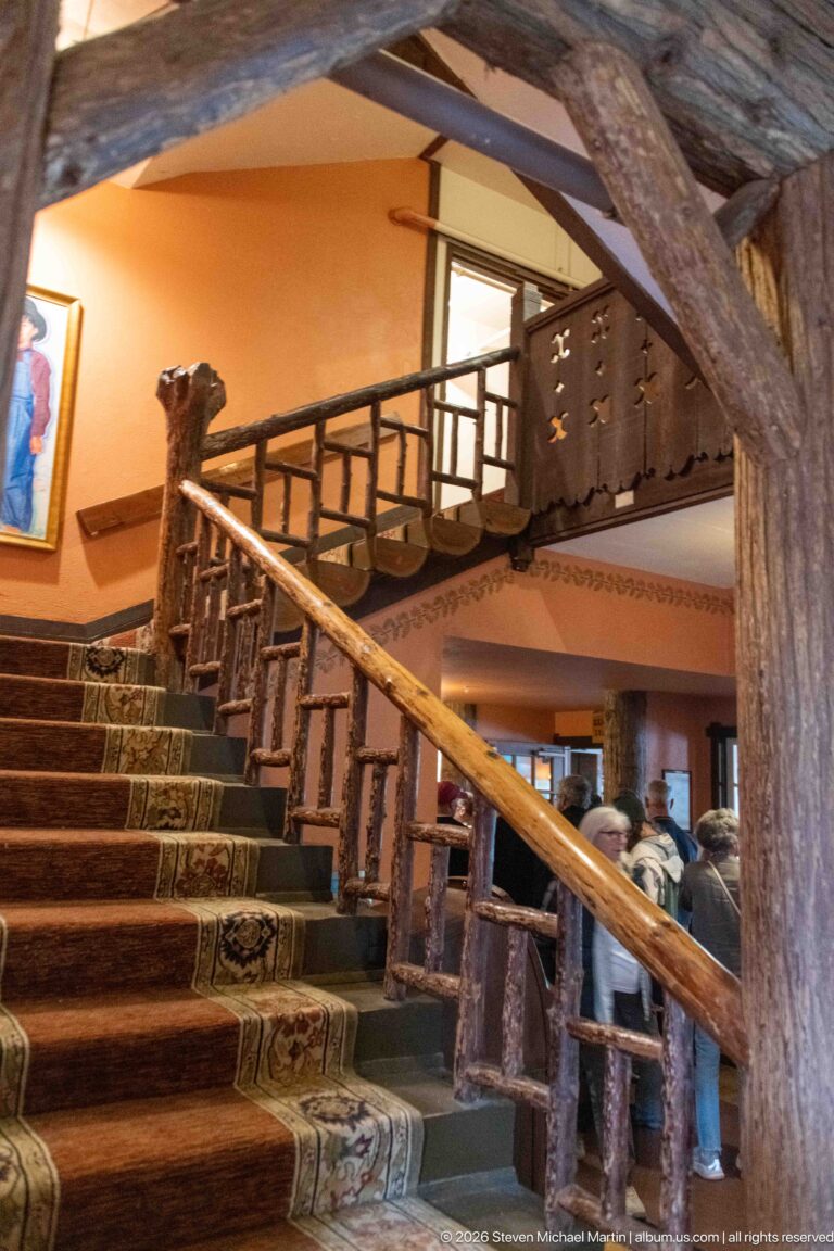 Broad carpeted staircase with log handrails, branch-form balusters, heavy beams, and an upper gallery inside Lake McDonald Lodge.