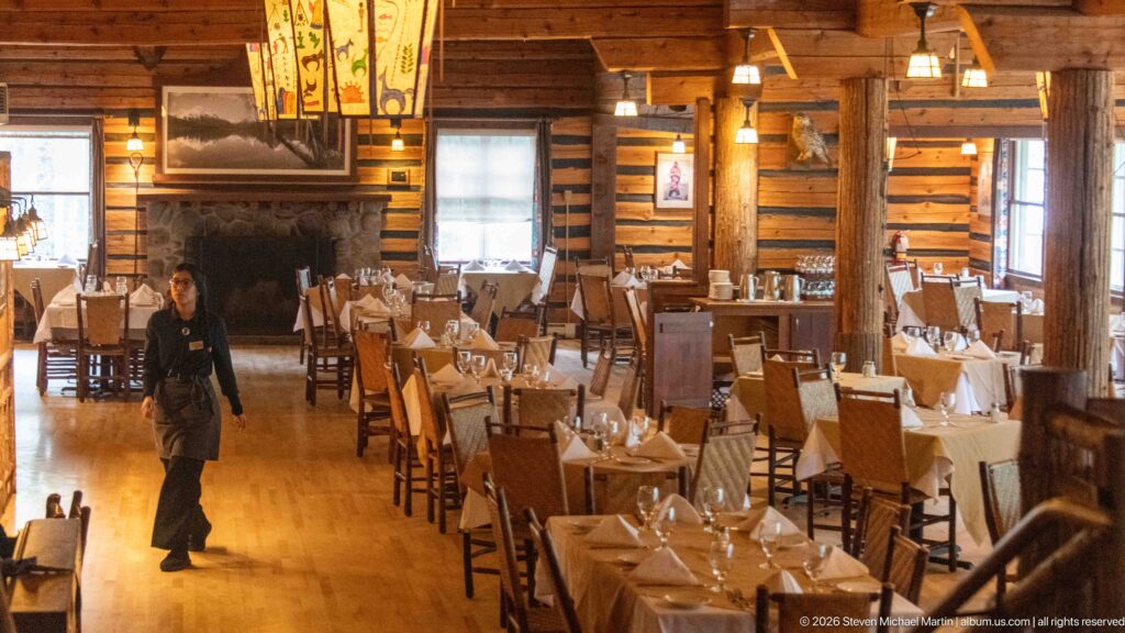 Lake McDonald Lodge dining room with set tables, log walls, timber columns, hanging lights, stone fireplace, and staff member.