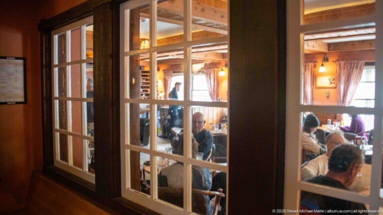 View through multi-pane glazed doors into a warmly lit Lake McDonald Lodge dining room with seated guests and tables.