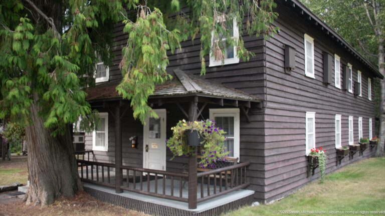 Corner view of a two-story dark wooden building with covered porch, white-trimmed windows, flowers, and a large tree.