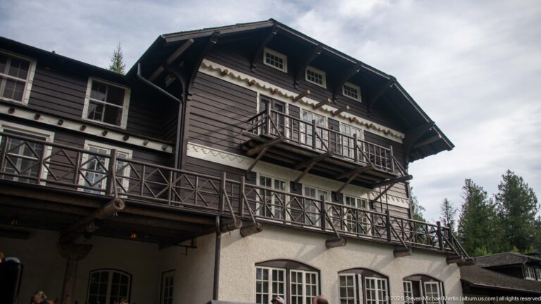 Close upward view of Lake McDonald Lodge with dark gabled upper story, projecting balconies, pale trim, and stucco lower level.