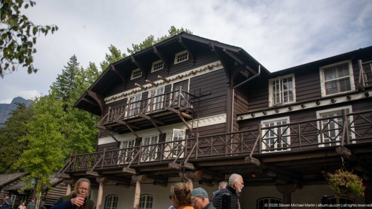 Upward exterior view of Lake McDonald Lodge central gable with dark siding, pale decorative bands, projecting balconies, and rustic railings.