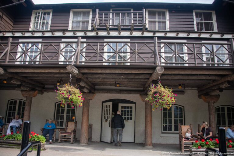 Frontal view of Lake McDonald Lodge gallery with log columns, cross-braced balcony railing, hanging flowers, windows, and visitors.