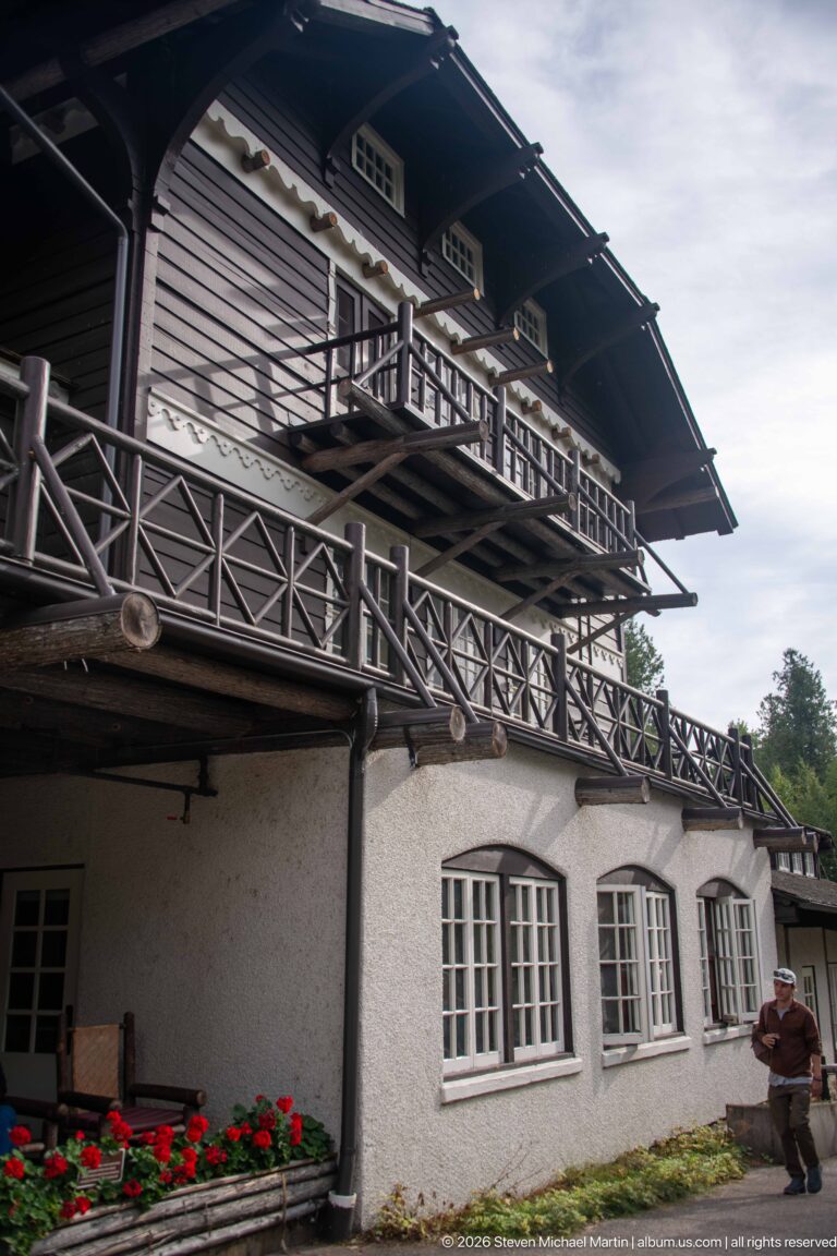 Angled exterior view of Lake McDonald Lodge showing a long dark timber balcony above pale stucco walls and multi-pane windows.