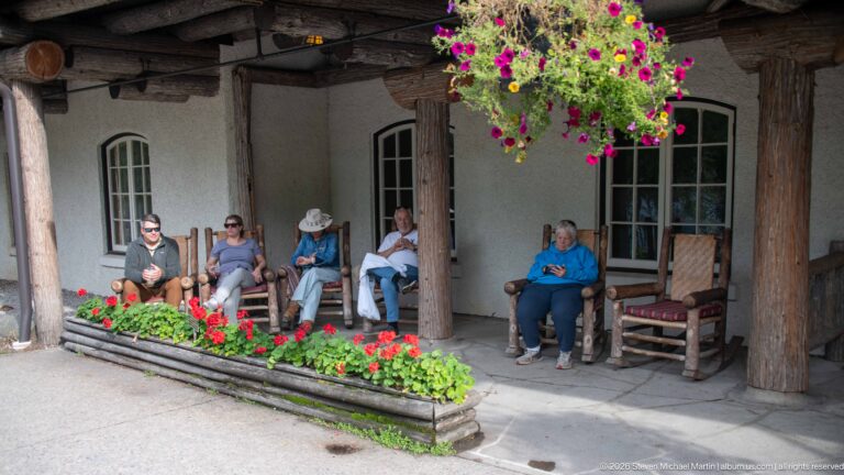 Visitors seated under a covered lodge gallery with thick log columns, hanging flowers, and a bright flower bed.
