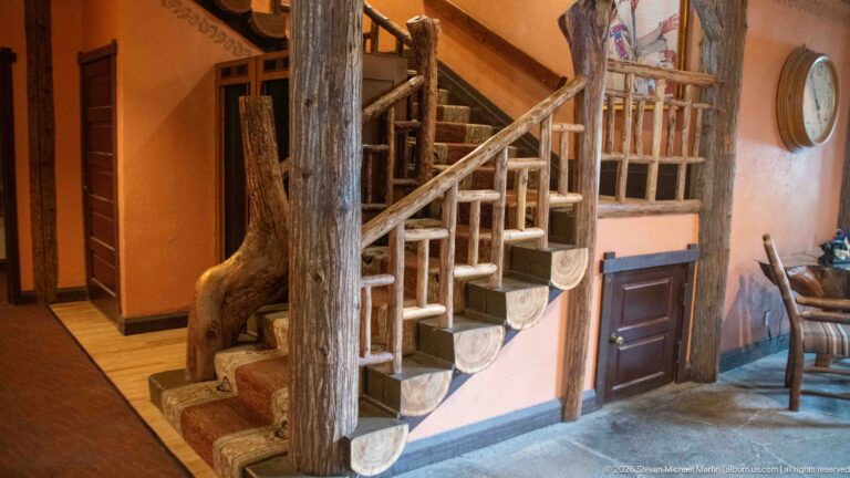 Interior staircase with carpeted steps, heavy log posts, natural-branch railings, and wood handrail at Lake McDonald Lodge.