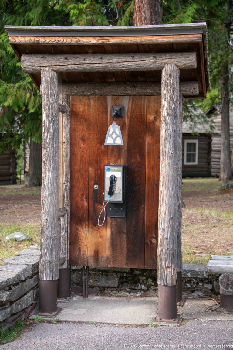 Wall-mounted public telephone beneath a small rustic shelter of heavy log posts, vertical wood boards, and a shallow roof.