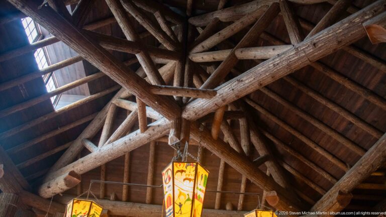 Upward view of exposed log roof trusses, diagonal braces, skylight, and hanging lanterns in Lake McDonald Lodge.