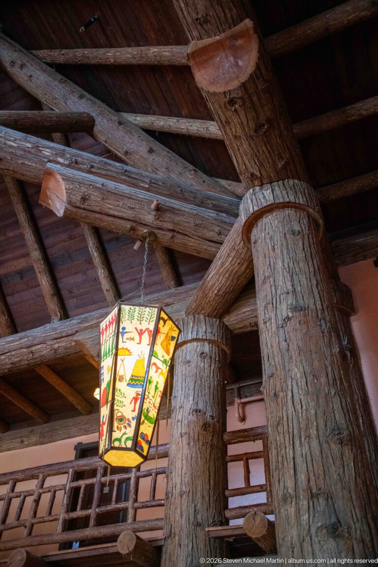 Upward view of massive log columns and beams with a painted hanging lantern and rustic balcony railing inside Lake McDonald Lodge.
