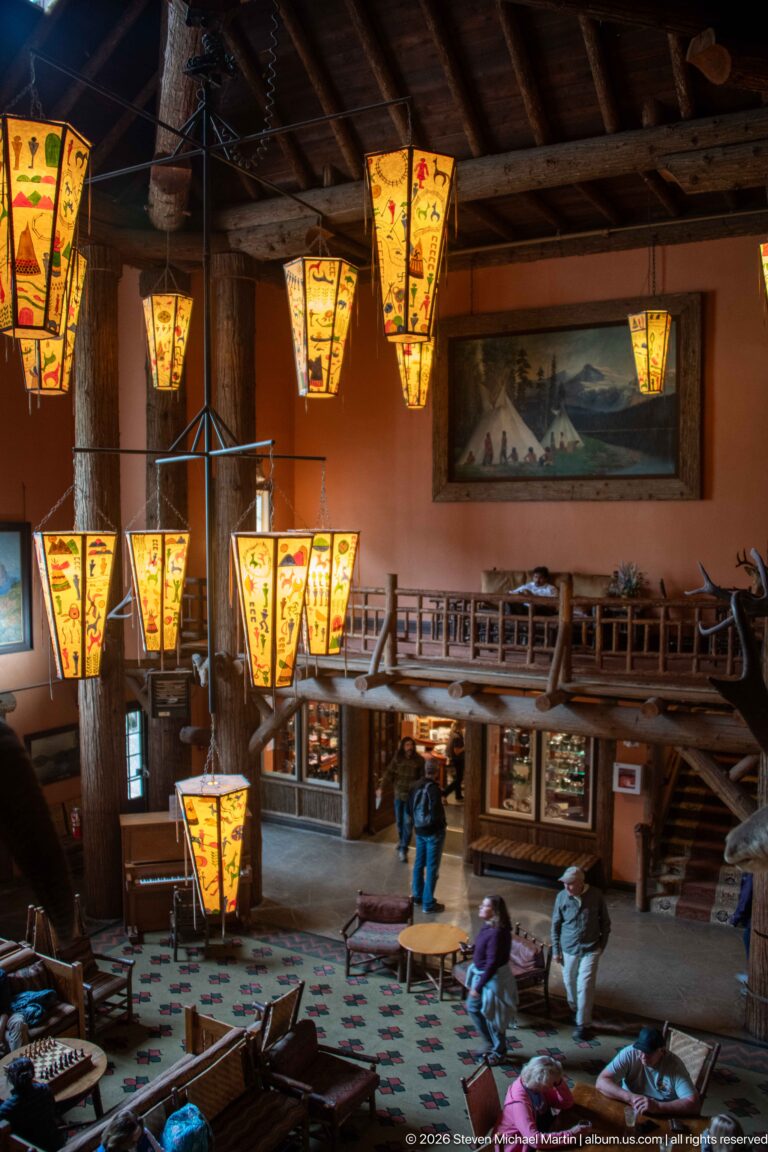 Lake McDonald Lodge lobby with painted hanging lanterns, massive log columns, mezzanine railings, fireplace, and seated visitors.