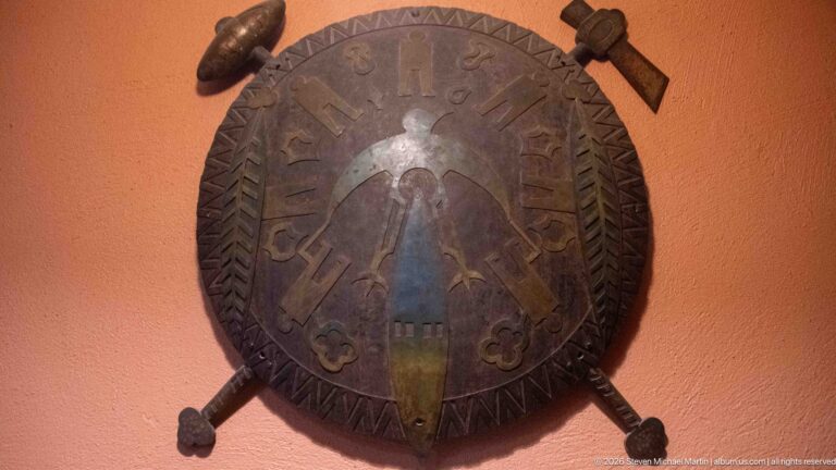 Round decorated wall hanging with geometric border, central painted figure, and crossed projecting elements on an orange wall.