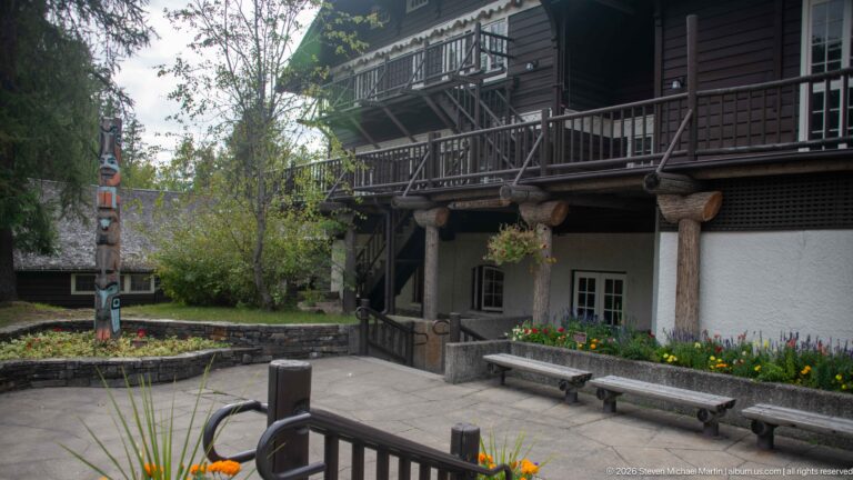 Close exterior view of Lake McDonald Lodge with log columns, dark timber balcony, pale walls, benches, flowers, and a carved post.