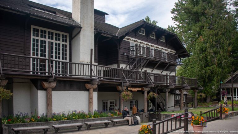 Multi-story Lake McDonald Lodge exterior with dark timber balconies, pale stucco walls, log supports, benches, and flower beds.