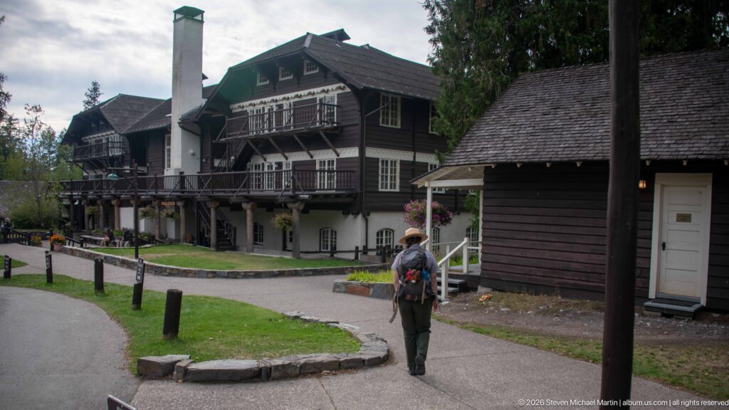 Visitor walking along a curved path between Lake McDonald Lodge and a small dark-sided outbuilding.