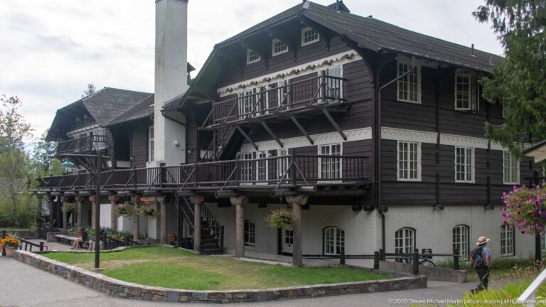 Multi-story Lake McDonald Lodge exterior with dark upper siding, white-framed windows, balconies, exterior stairs, and tall chimney.