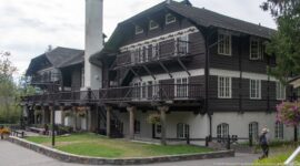 Multi-story Lake McDonald Lodge exterior with dark upper siding, white-framed windows, balconies, exterior stairs, and tall chimney.