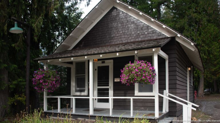 Small dark wooden building with steep shingled gable, white porch railings, entry door, ramp, and hanging purple flowers.