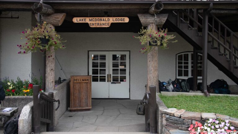 Covered lodge entrance with white double doors, log posts, hanging flowers, bellman stand, exterior stair, and entrance sign.