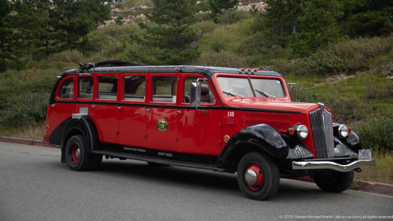 Historic red Glacier National Park touring bus in side view, with black fenders, chrome grille, and roll-back roof.