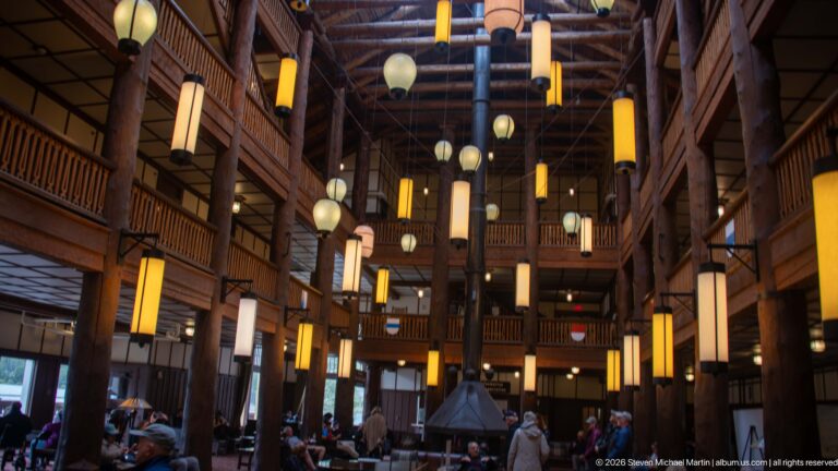 Many Glacier Hotel Great Room with hanging lanterns, massive log columns, stacked balconies, central fireplace, and guests.