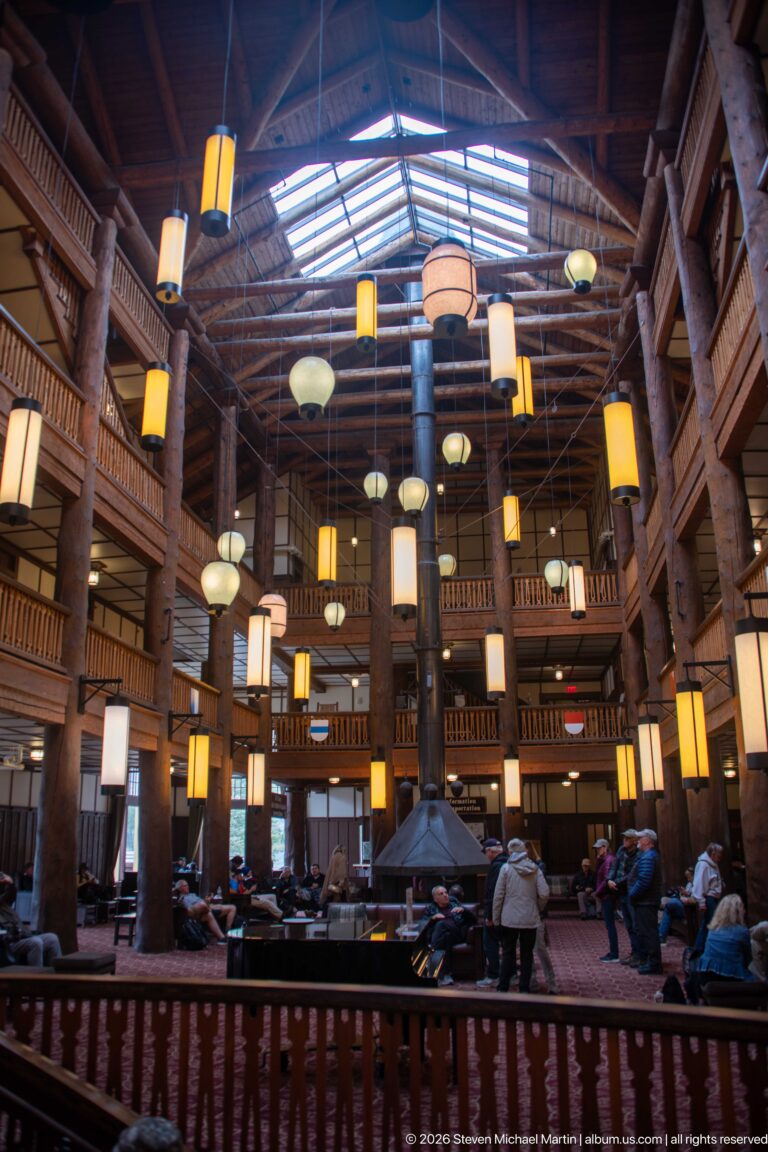 Wide Great Room interior with log columns, multi-level balconies, hanging lanterns, central fireplace, and visitors.