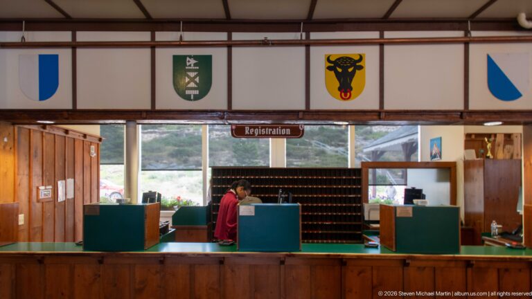 Front-facing registration counter with green stations, staff member, key-slot wall, and heraldic shields above.